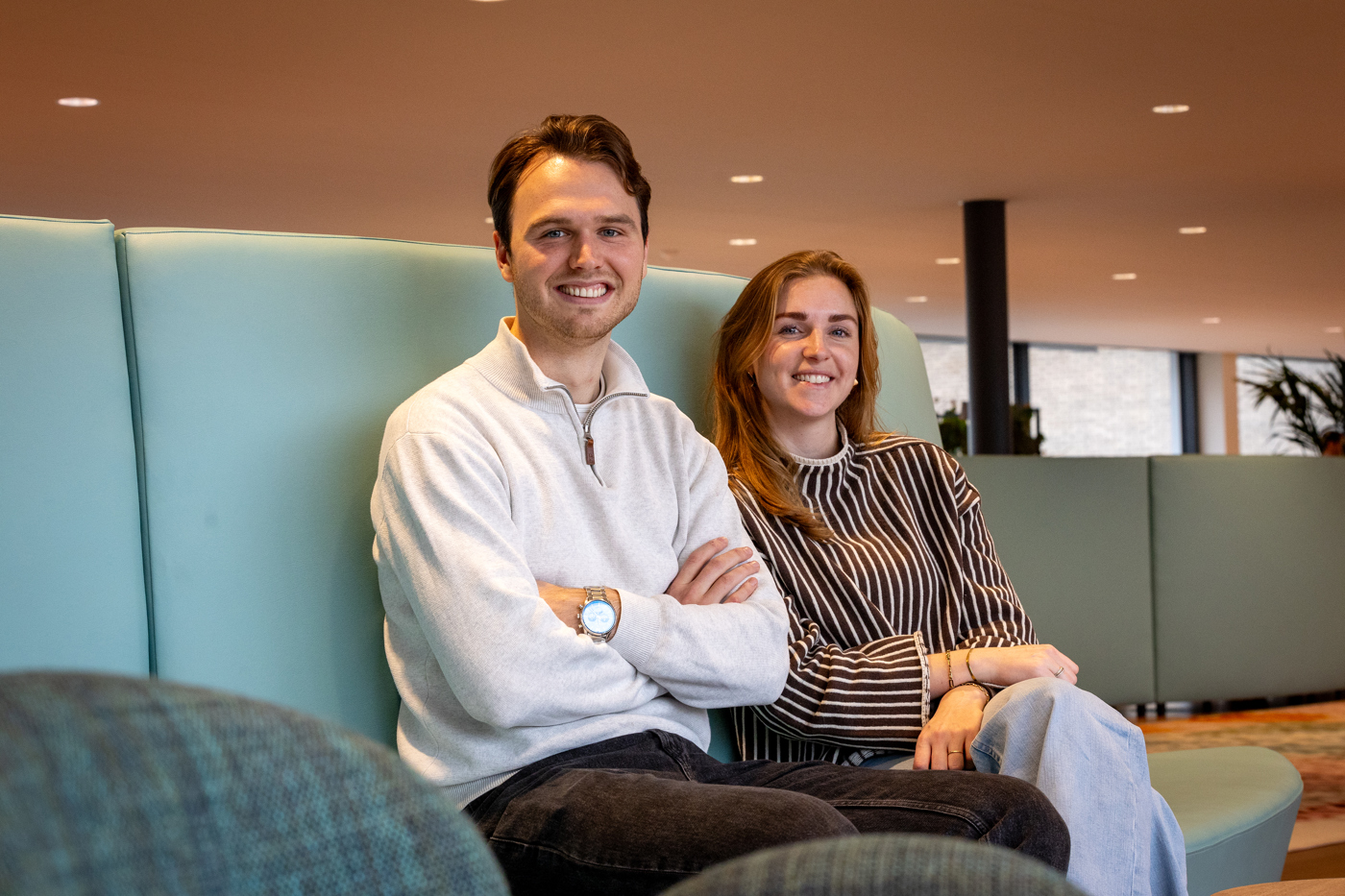 Collega's Nick en Laura zitten naast elkaar op een blauwe bank. Collega Nick draagt een gruize trui en een zwarte broek. Collega Laura draagt een zwart wit gestreepte trui en een spijkerbroek.
