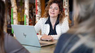 Collega Marijke zit achter een laptop aan een tafel. ze draagt een wit vest. Op de voorgrond zitten nog twee andere collega's. 