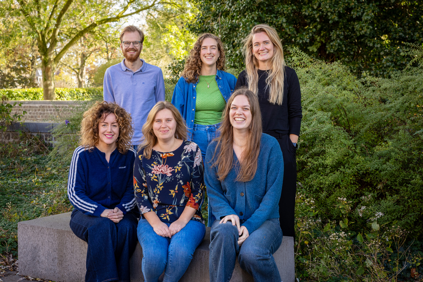 Collega's van team participatie zitten op en staan achter een bankje buiten het provinciehuis. Drie vrouwelijke collega's zitten op de bank en de andere drie collega's, twee vrouwen en een man staan achter de zitten collega's. Er is veel groen op de achtergrond.
