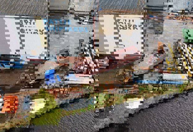 Op de eerste afbeelding is een vrachtwagen te zien die het casco van het werkschip een loods binnen rijdt. Op de tweede afbeelding is met een drone buiten de loods waar het werkschip gemaakt wordt, van bovenaf een foto gemaakt van het casco.