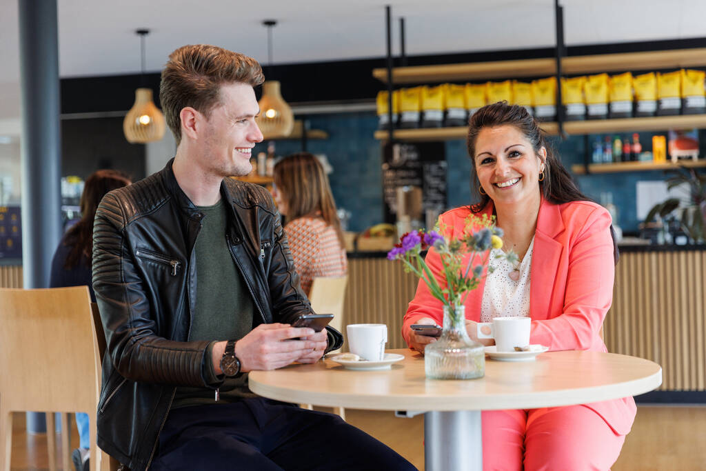 2 collega's zitten aan een ronde tafel. De vrouw draagt een roze pak en de man en zwarte broek met een groen shirt en een zwart jasje.