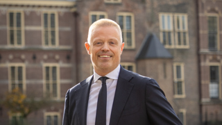 Collega Paul staat tegen een reling aan bij de Hofvijver in Den Haag. Hij draagt een donkerblauw pak.