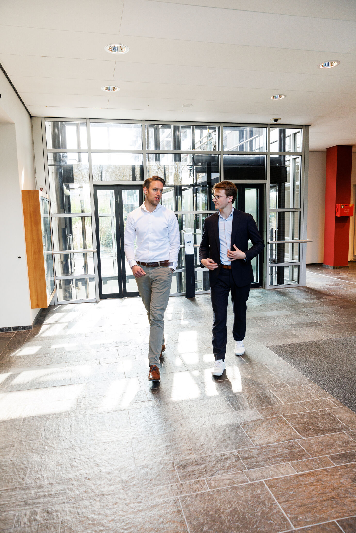 Twee jonge mannen in pak lopen richting de camera. Ze zijn in gesprek met elkaar.