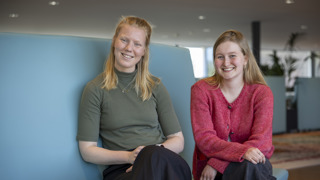 Lidewij en Mirthe zitten op een blauwe bank naast elkaar en kijken in de camera.