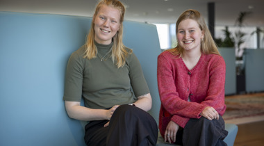 Lidewij en Mirthe zitten op een blauwe bank naast elkaar en kijken in de camera.
