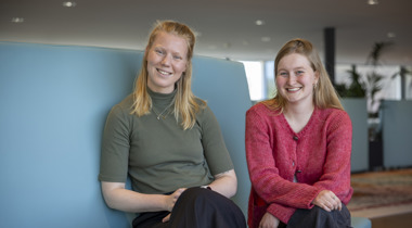 Lidewij en Mirthe zitten op een blauwe bank naast elkaar en kijken in de camera.