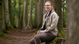 Collega Annemarie zit tussen bomen op een boomstronk. Ze draagt bruinte kleding en heeft een verrekijker om haar nek hangen.
