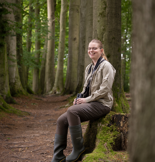 Collega Annemarie zit tussen bomen op een boomstronk. Ze draagt bruinte kleding en heeft een verrekijker om haar nek hangen.