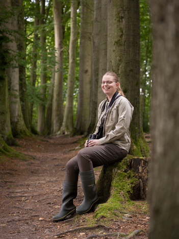 Collega Annemarie zit tussen bomen op een boomstronk. Ze draagt bruinte kleding en heeft een verrekijker om haar nek hangen. Op de tweede afbeelding zit collega Annemarie gehurkt tussen het groen en paarse bloemen in een weide.