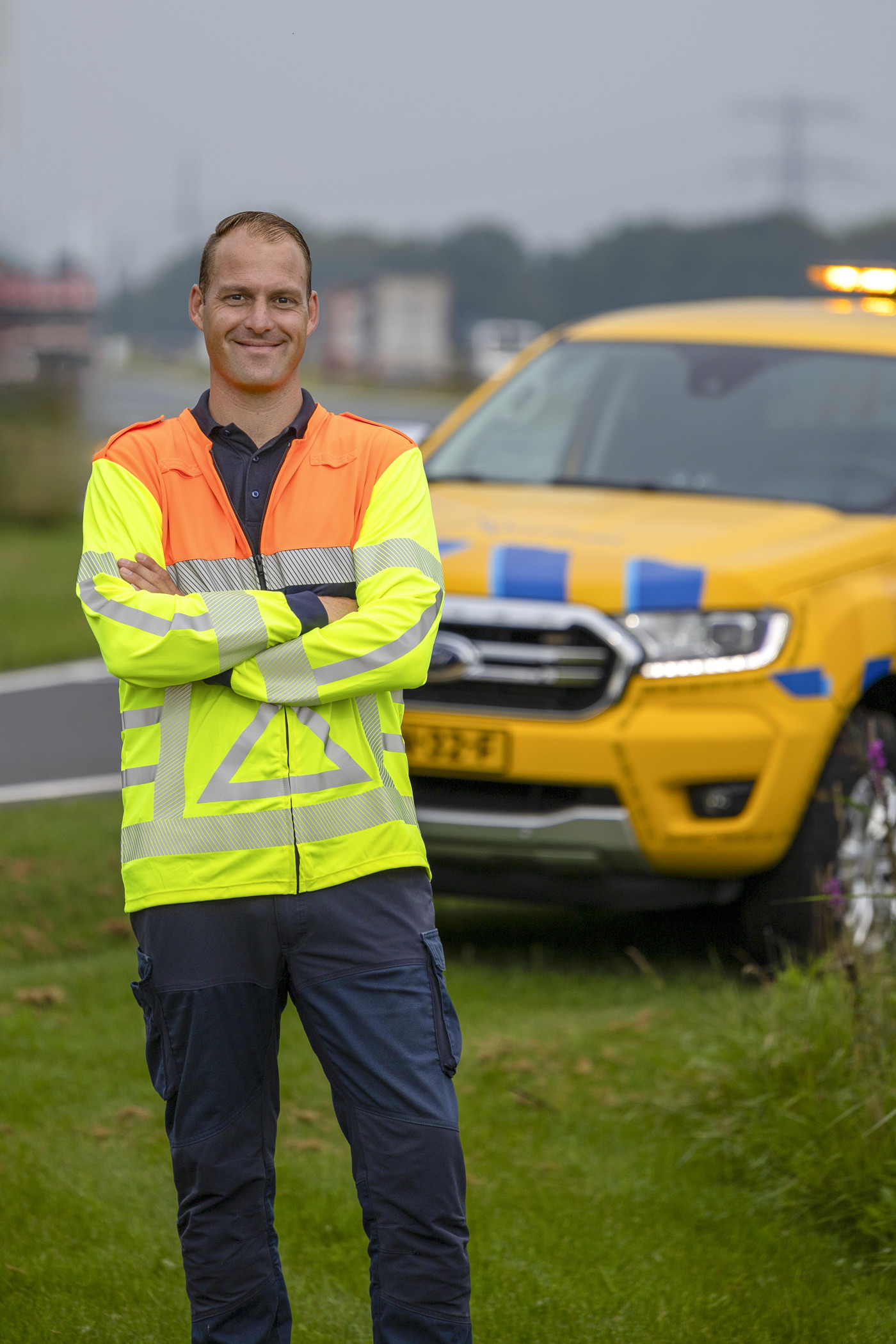 Collega Luuk staat met z'n armen over elkaar voor een geel blauwe provincie auto. Hij staat aan de zijkant van een weg, bij de berm Hij draagt een oranje geel hesje.