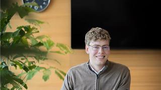 Collega Rudmer zit op een witte stoel aan een bruine tafel en werkt achter zijn laptop terwijl hij naar de camera kijkt.