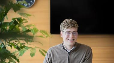 Collega Rudmer zit op een witte stoel aan een bruine tafel en werkt achter zijn laptop terwijl hij naar de camera kijkt.