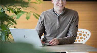 Collega Rudmer zit op een witte stoel aan een bruine tafel en werkt achter zijn laptop terwijl hij naar de camera kijkt.