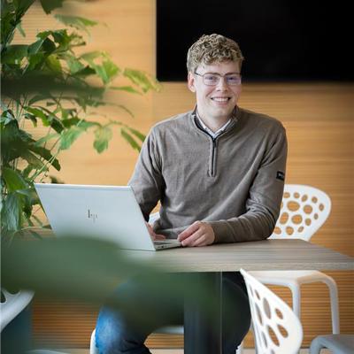 Collega Rudmer zit op een witte stoel aan een bruine tafel en werkt achter zijn laptop terwijl hij naar de camera kijkt.
