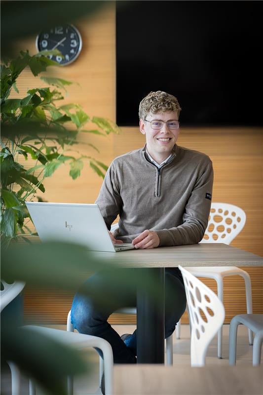 Collega Rudmer zit op een witte stoel aan een bruine tafel en werkt achter zijn laptop terwijl hij naar de camera kijkt.
