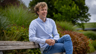Trainee Wouter zit op een bankje met veel groen op de achtergrond. Hij draagt een spijkerbroek en een blauw wit gestreept overhemd.