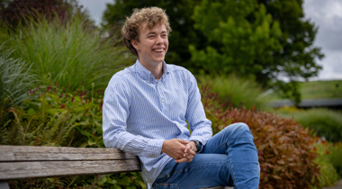 Trainee Wouter zit op een bankje met veel groen op de achtergrond. Hij draagt een spijkerbroek en een blauw wit gestreept overhemd.