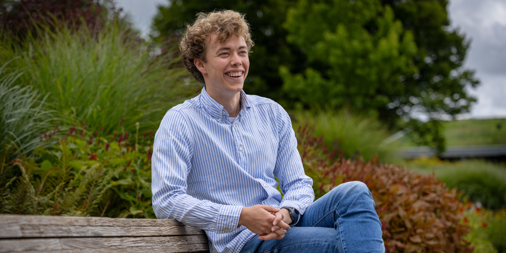 Trainee Wouter zit op een bankje met veel groen op de achtergrond. Hij draagt een spijkerbroek en een blauw wit gestreept overhemd.