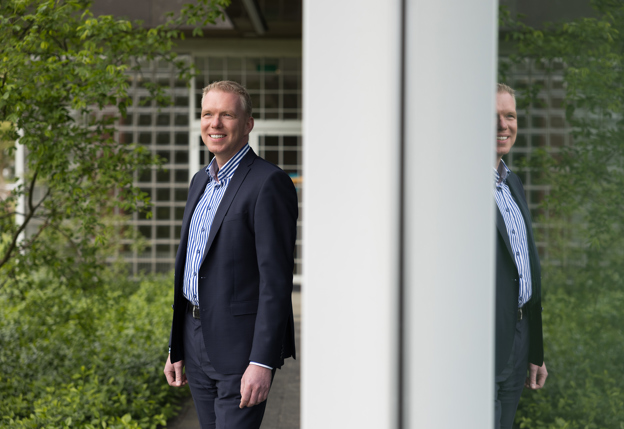 Op de eerste afbeelding is collega Henri te zien naast een raam buiten het provinciehuis. Hij is in de weerspiegeling nog een keer te zien. Op de tweede afbeelding is collega Melanie te zien, ze zit op een blauwe bank aan een tafel en werkt acher een laptop.