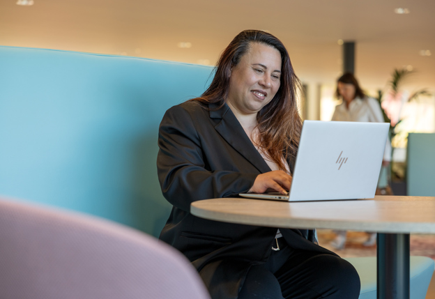 Op de eerste afbeelding is collega Henri te zien naast een raam buiten het provinciehuis. Hij is in de weerspiegeling nog een keer te zien. Op de tweede afbeelding is collega Melanie te zien, ze zit op een blauwe bank aan een tafel en werkt acher een laptop.