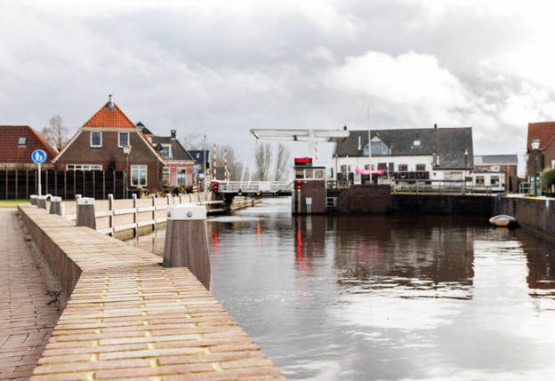 Op de eerste afbeelding is een kade in Ossenzijl te zien en op de tweede afbeelding staat diezelfde kade op de foto met collega's Johan en Bart erbij. Collega Johan wijst naar het water en is wat door zijn knieën.