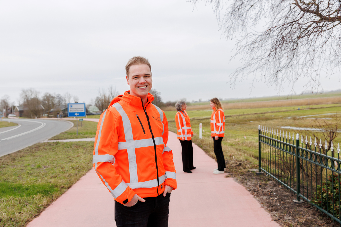 Op de voorgrond van de afbeelding staat collega Tiemen in een oranje hesje van de provincie. Op de achtergrond staan collega's Maud en Eiberlien en zijn met elkaar in gesprek.