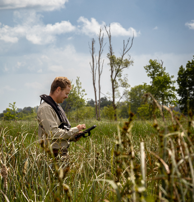 Collega Rinze staat in een natuur gebied in een veld met op de achtergrond wat bomen. Hij kijkt op een tablet.