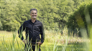 Collega Ives staat in een weiland met op de achtergrond een bos. Hij draagt een zwarte blouse en een zwarte broek.