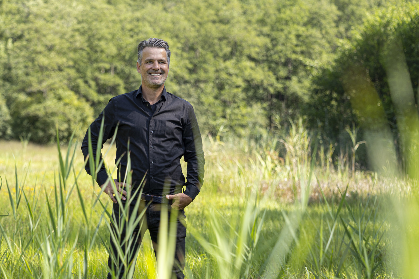 Collega Ives staat in een weiland met op de achtergrond een bos. Hij draagt een zwarte blouse en een zwarte broek.