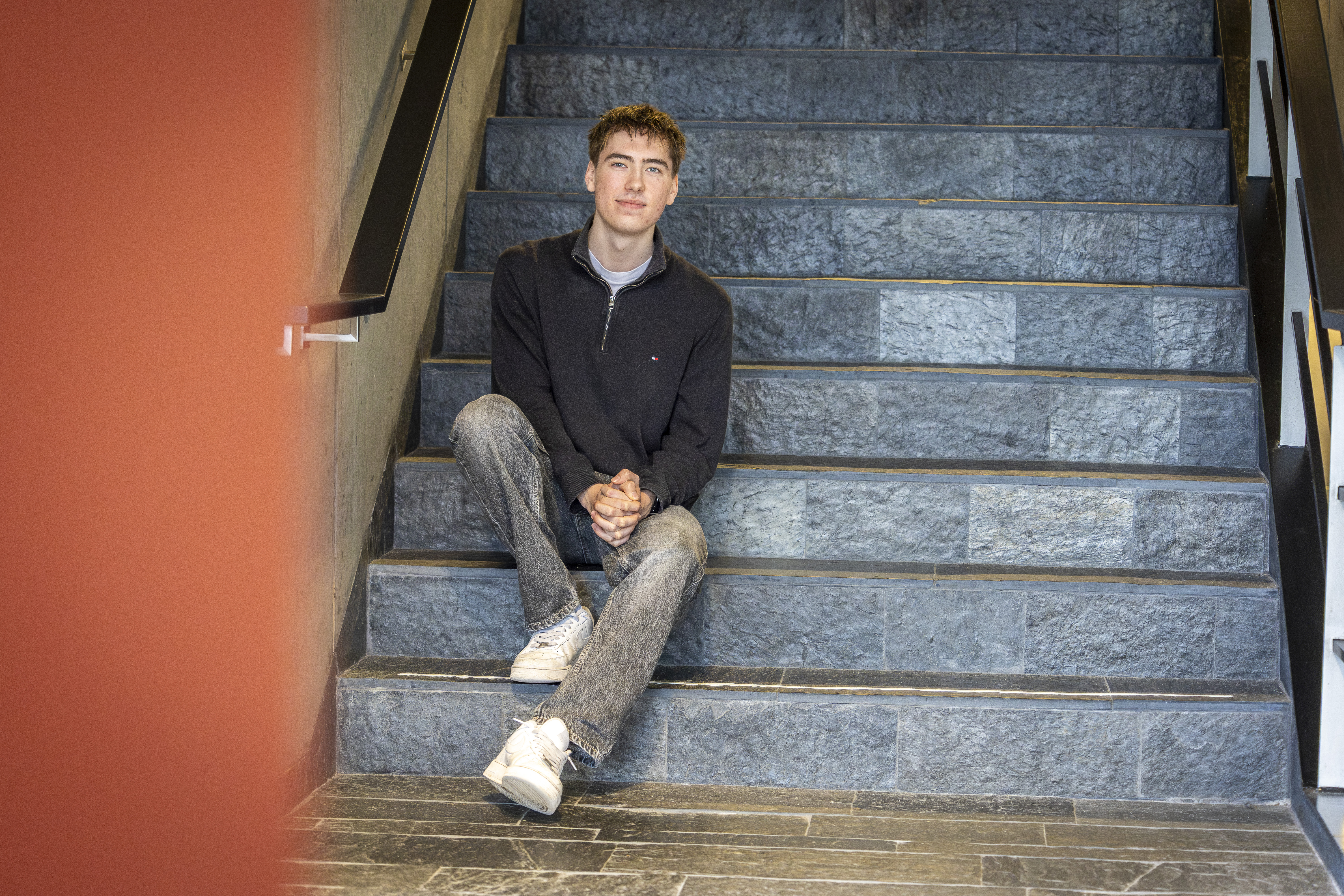 Stagiair San Mei zit op een trap. Hij draagt een grijze spijkerbroek en een blauwe trui.