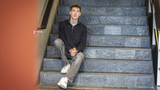 Stagiair San Mei zit op een trap. Hij draagt een grijze spijkerbroek en een blauwe trui.