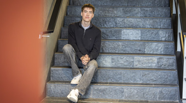 Stagiair San Mei zit op een trap. Hij draagt een grijze spijkerbroek en een blauwe trui.