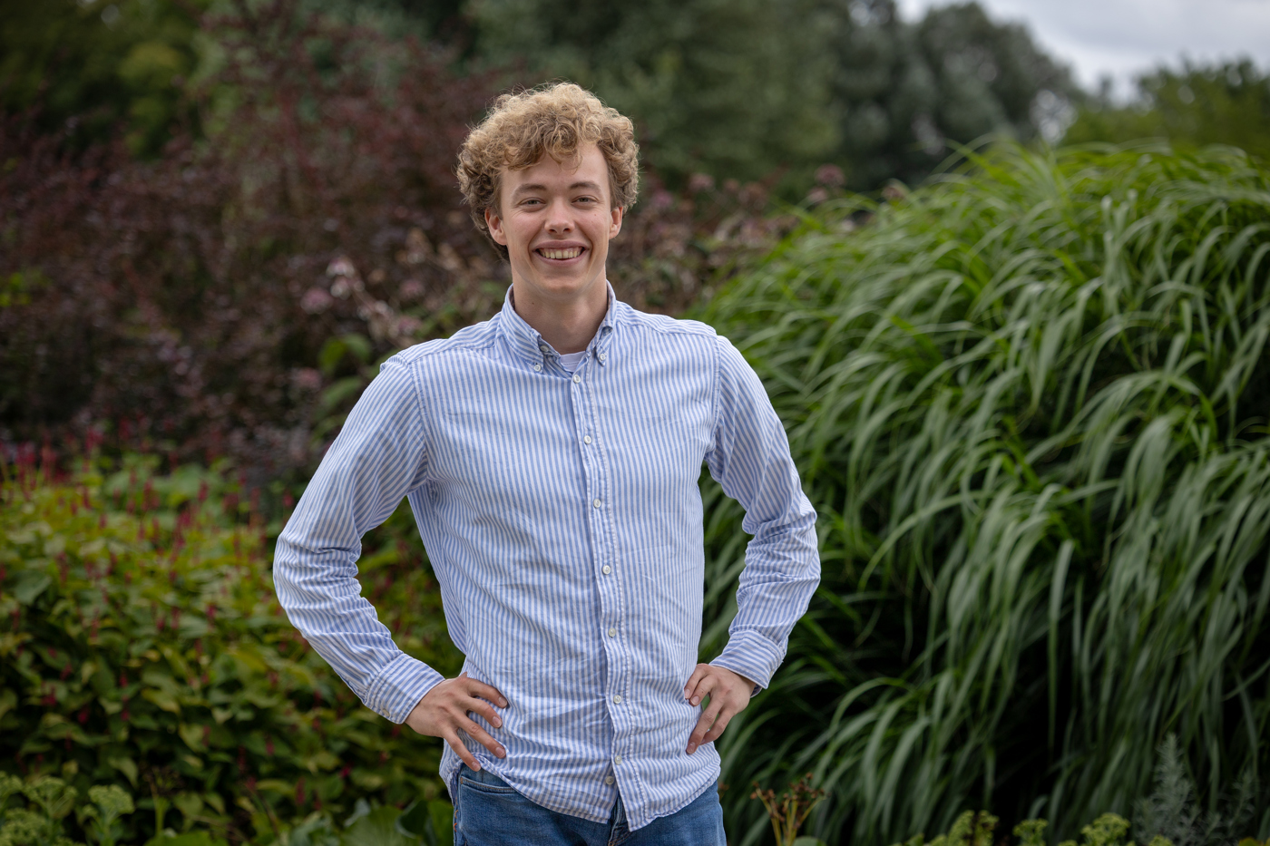 Trainee Wouter draagt een blauw wit gestreept overhemd en een spijkerbroek. Hij staat voor een groene omgeving met planten en struiken.