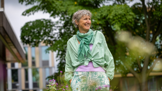 Collega Dicky staat tussen paarse bloemen in de tuin bij het provinciehuis. Op de achtergrond is een boom te zien. Dicky draagt een groene blouse en een paars shirt. Daaronder een wit met groene broek.