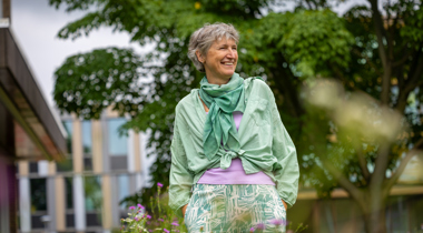 Collega Dicky staat tussen paarse bloemen in de tuin bij het provinciehuis. Op de achtergrond is een boom te zien. Dicky draagt een groene blouse en een paars shirt. Daaronder een wit met groene broek.
