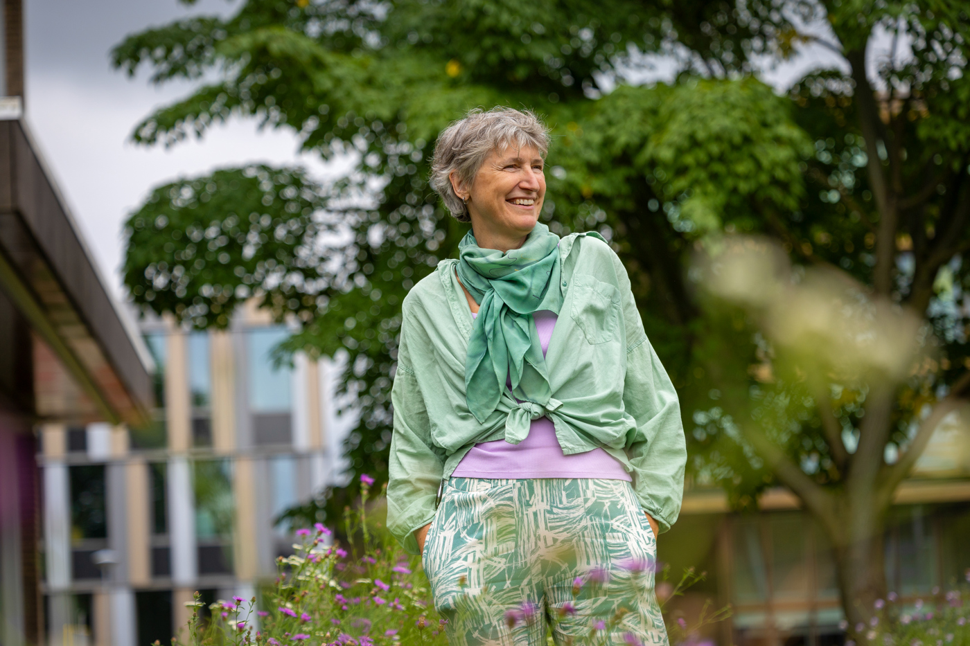 Collega Dicky staat tussen paarse bloemen in de tuin bij het provinciehuis. Op de achtergrond is een boom te zien. Dicky draagt een groene blouse en een paars shirt. Daaronder een wit met groene broek.