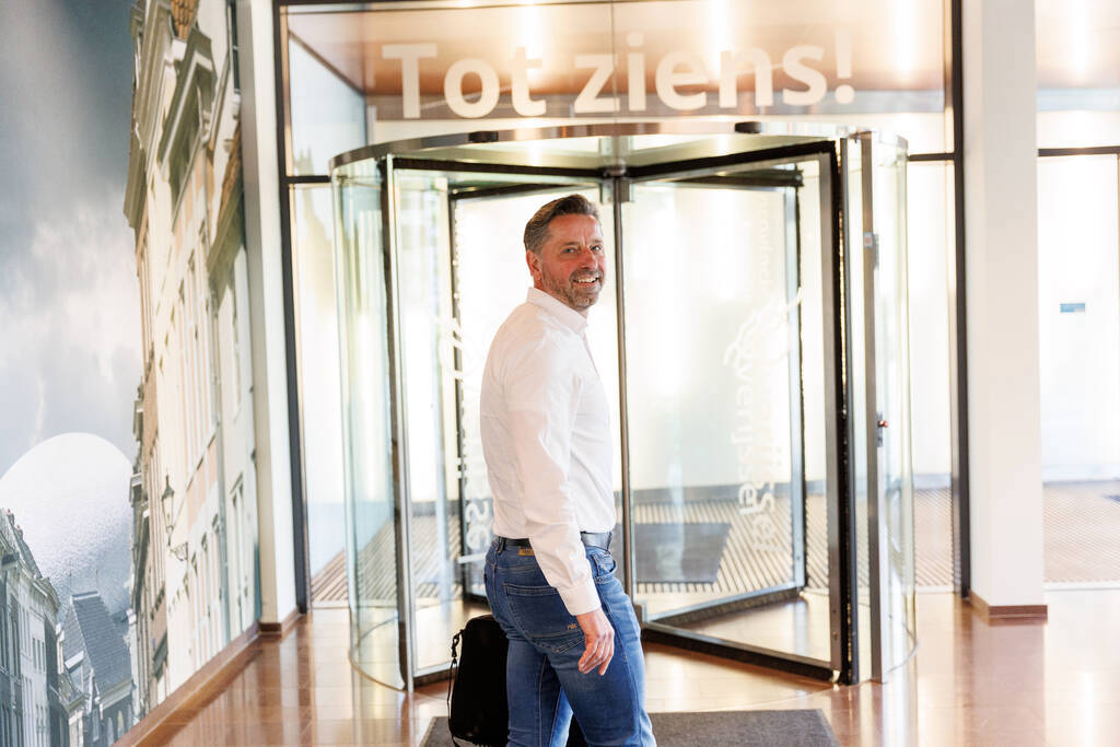 Op de afbeelding is collega Bas te zien, die binnen voor de draaideuren van de in- en uitgang van het provinciehuis van Overijssel staat. Boven de draaideuren staan de woorden tot ziens.