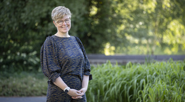Collega Marieke staat buiten het provinciehuis in de tuin. Op de achtergrond zijn grassen en bomen te zien. Marieke draagt een bril en heeft kort blond haar.