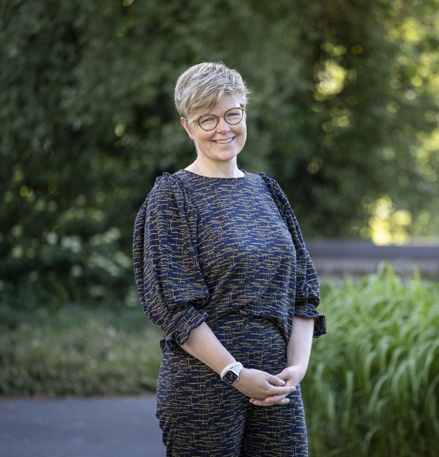 Collega Marieke staat buiten het provinciehuis in de tuin. Op de achtergrond zijn grassen en bomen te zien. Marieke draagt een bril en heeft kort blond haar.