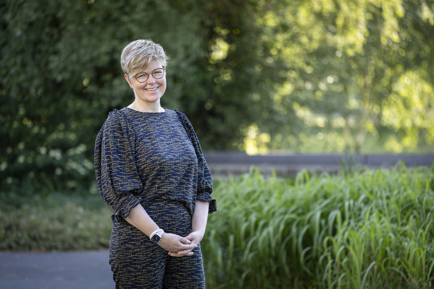 Collega Marieke staat buiten het provinciehuis in de tuin. Op de achtergrond zijn grassen en bomen te zien. Marieke draagt een bril en heeft kort blond haar.