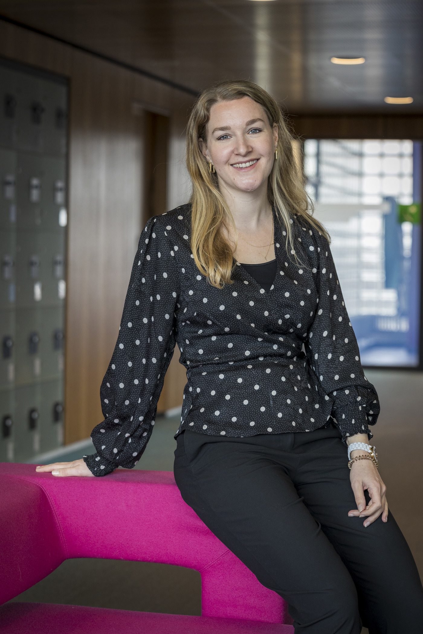 Collega Miriam zit op de leuning van een roze bank en draagt en zwarte blouse met witte stippen.
