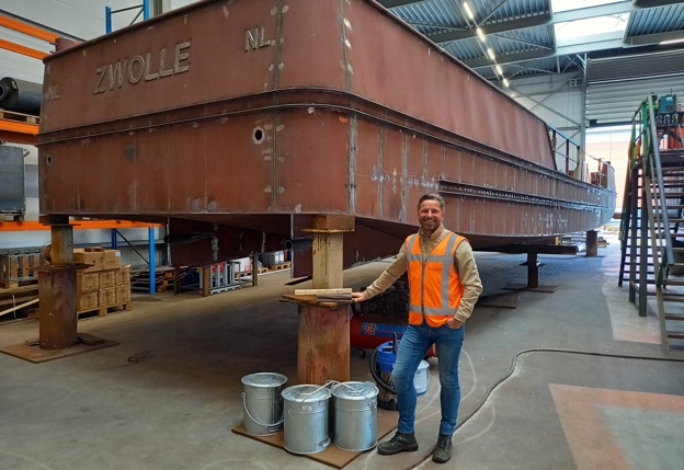 Op de eerste afbeelding is collega Bas Spliet te zien die naast het casco van het werkschip staat. Op de tweede foto is de nieuwe casco te zien van het werkschip in de scheepswerf van Groenveldt Marine Contruction in Hendrik-Ido-Ambacht.