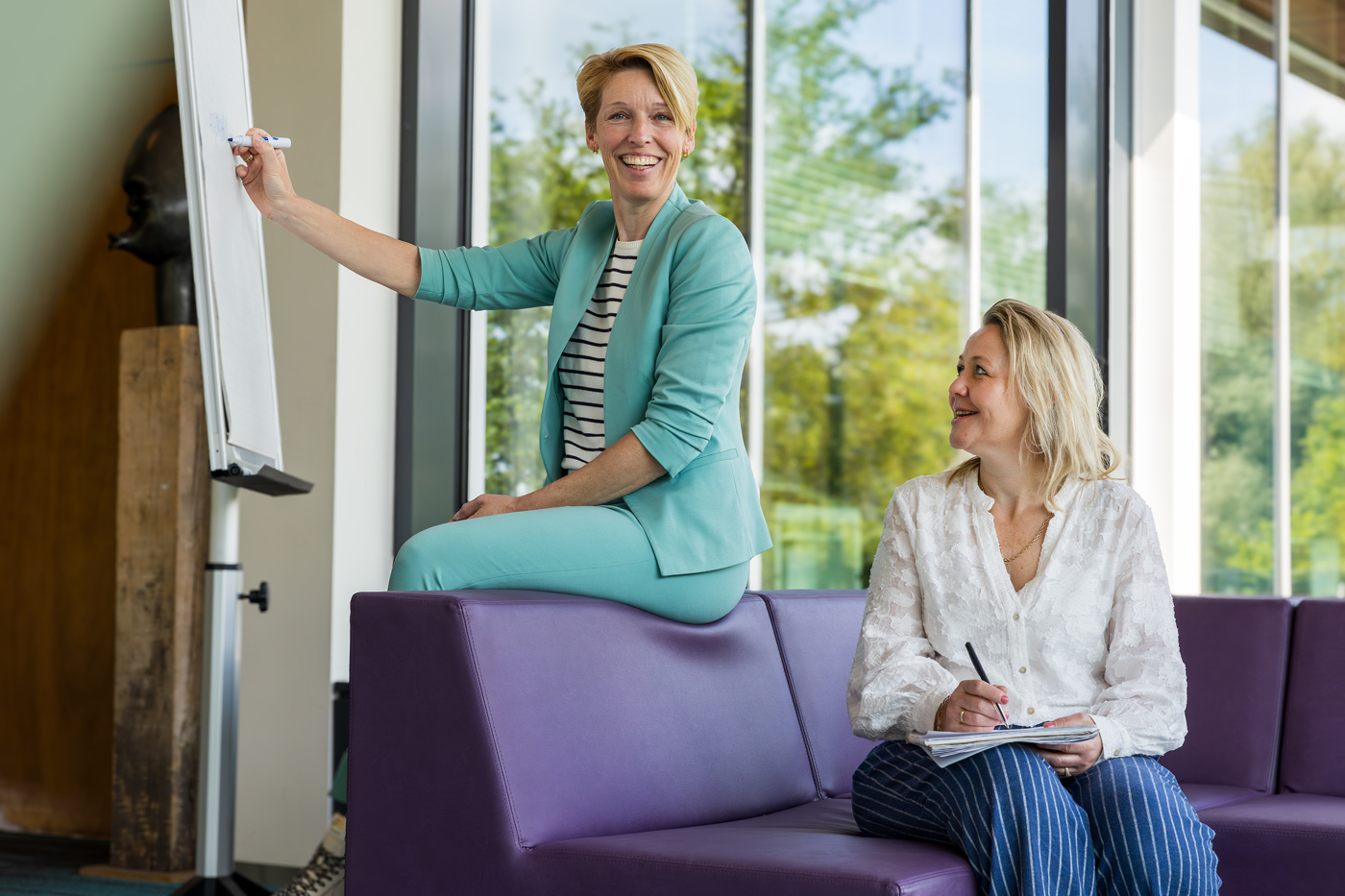 Collega's Linda en Barbara zijn te zien op de foto. Linda zit op de leuning van de paarse bank en schrijft iets op een groot vel papier en Barbara zit op de paarse bank naar haar te kijken met pen en papier in haar hand.