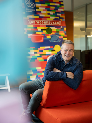 Op de eeste afbeelding zit collega Björn op een rode bank en achter hem is een plaat te zien met veel verschillende kleuren en de titel woonkeuken staat erop. Op de tweede afbeelding staat collega Marieke in de tuin buiten het provinciehuis. Er zijn bomen en grassen te zien op de achtergrond.