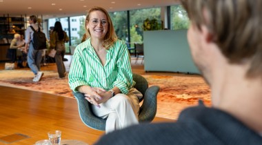 Collega Lyanne is in gesprek met een andere collega. Ze draagt een groene blouse en een witte broek. Ze zit op een blauwe stoel.