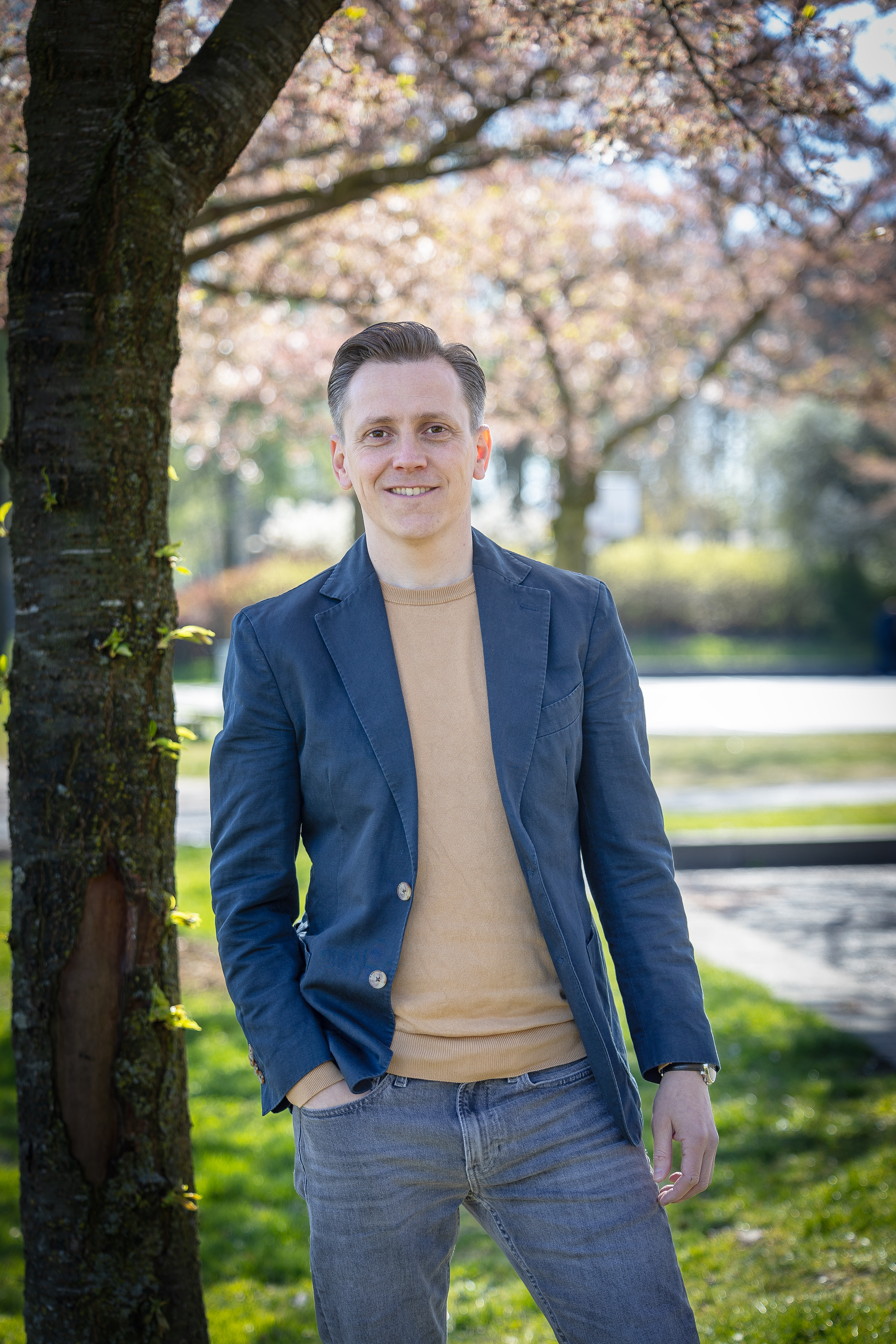 Collega Robbert staat buiten het provinciehuis op het gras naast een boom. Hij heeft een spijkerbroek aan en heeft zijn rechterhand in zijn broekzak. Hij draagt een blauw jasje en beige trui.