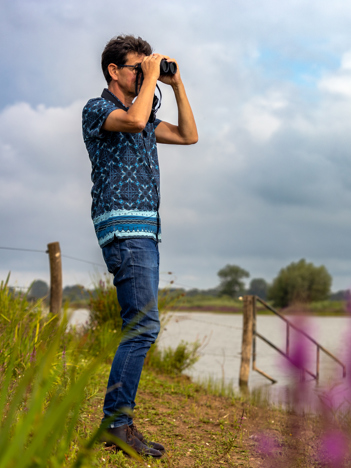 Collega Chris zit gehurkt tussen de natuur in het Vreugderijkerwaard op de eerste afbeelding. Collega Chris kijkt door een verrekijker bij het Vreugderijkerwaard op de tweede afbeelding.